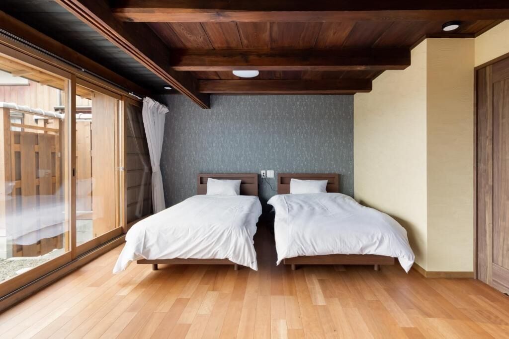 A room with two beds, wooden flooring, a sloped ceiling, and a large window with curtains.