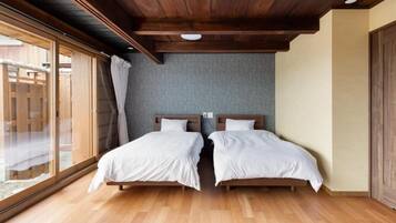 A room with two beds, wooden flooring, a sloped ceiling, and a large window with curtains.