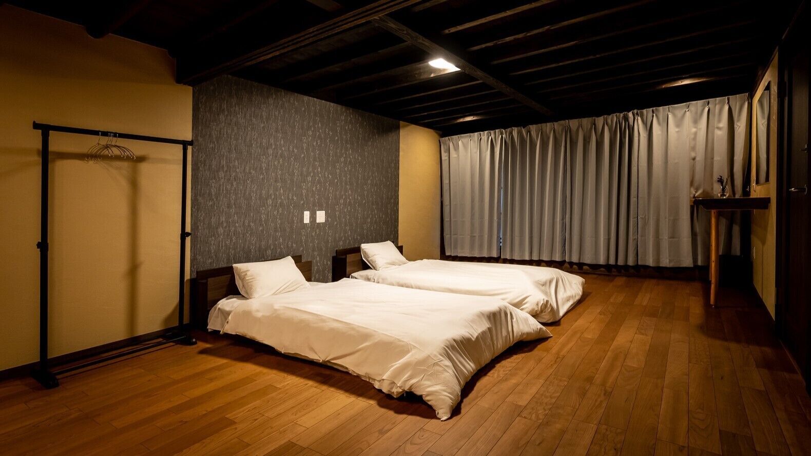 Una habitación con dos camas, suelo de madera, pared oscura con textura y una ventana cubierta por cortinas.