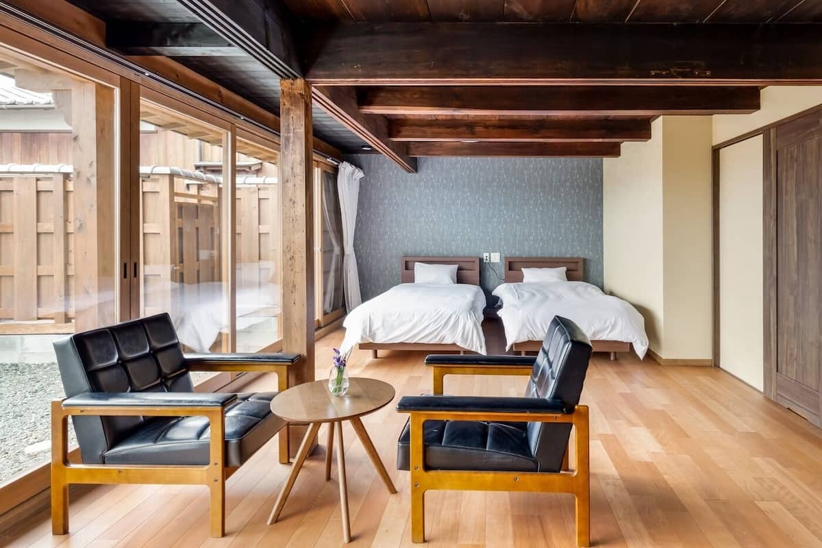 Una camera d'albergo con due letti, soffitto in legno, un'ampia finestra e un piccolo tavolo rotondo con sedie.