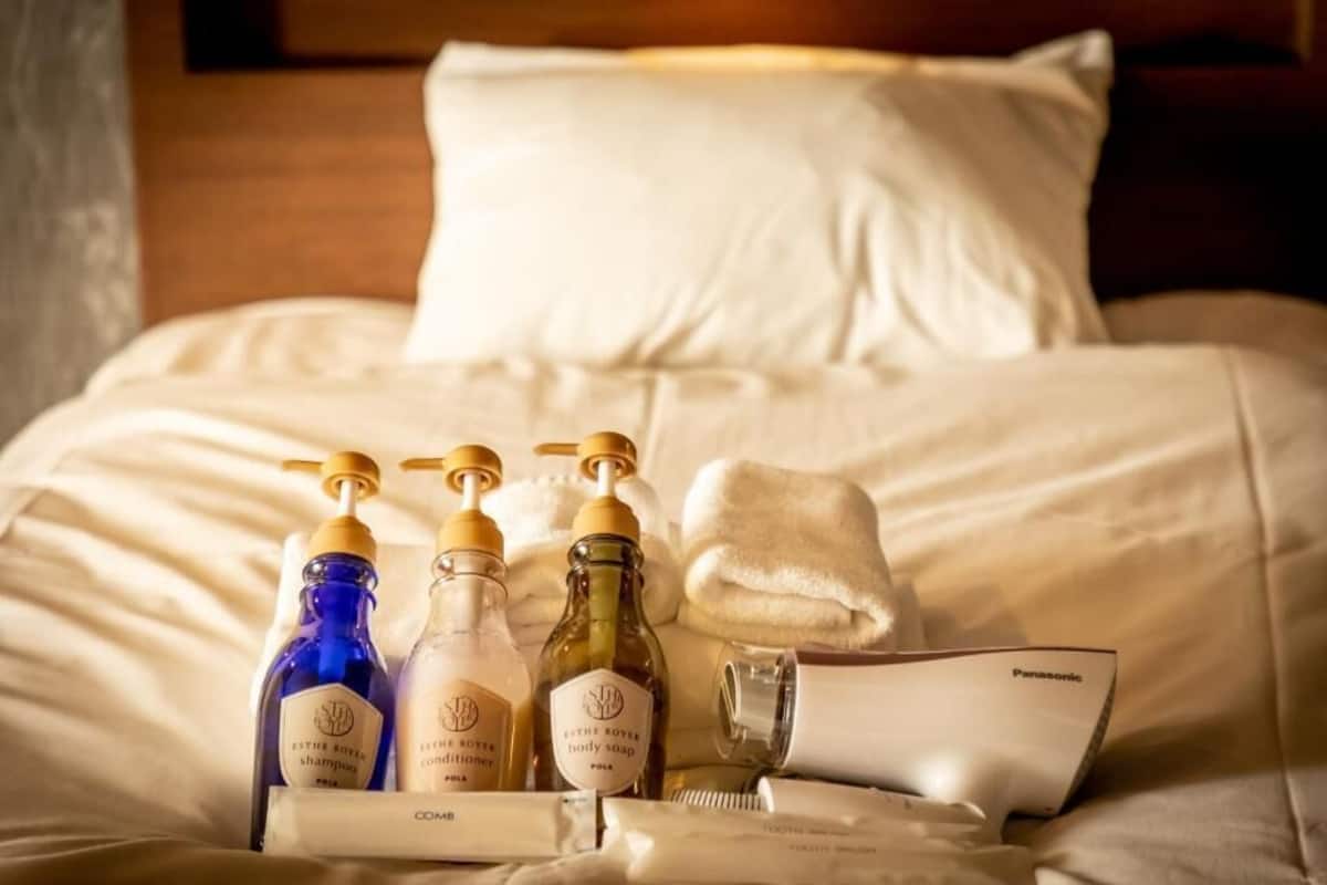 Un set di articoli da toilette ordinato su un letto con lenzuola bianche, tra cui shampoo, balsamo, sapone corpo e uno spazzolino da denti.