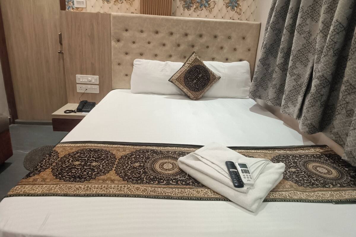 A neatly made bed with a patterned bedspread, a single decorative pillow, and a folded towel. There is a bedside table with a phone and a remote control. The room has floral wallpaper and a wooden door.