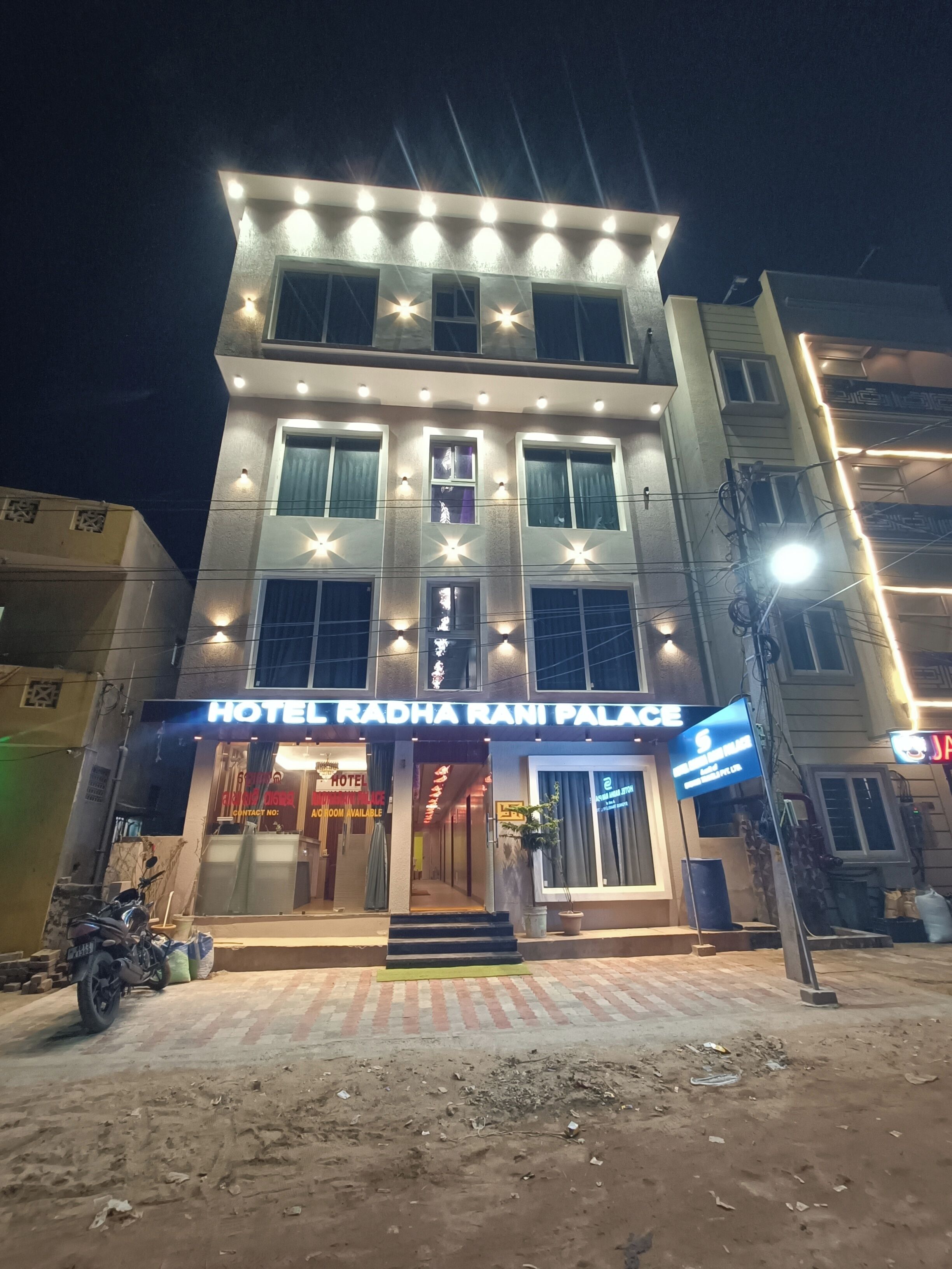 Un hotel llamado "Hotel Radha Rani Palace" con varios pisos, ventanas iluminadas y una entrada visible.