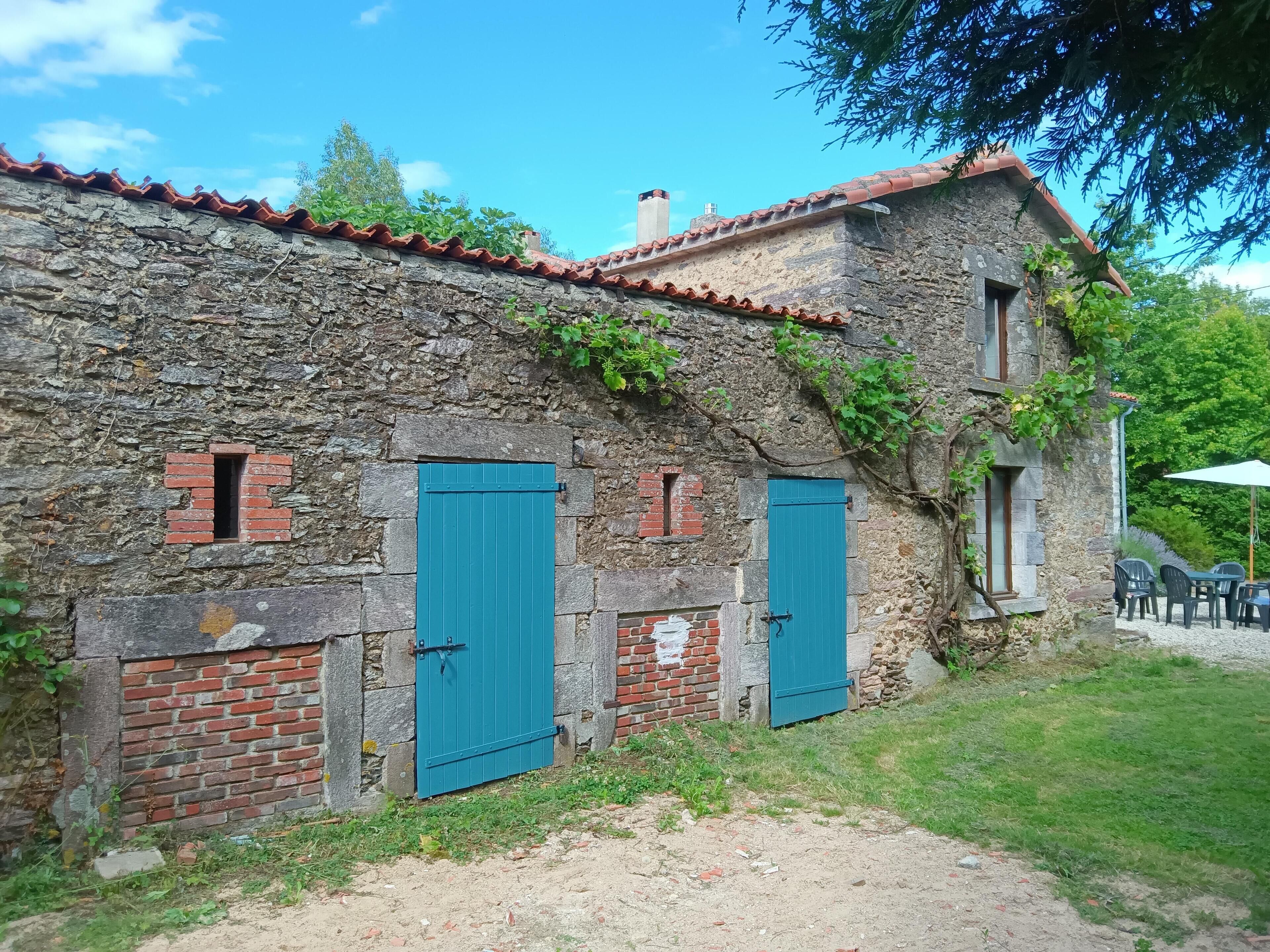 Un edificio rústico de piedra con dos puertas azules y un ventanal pequeño, rodeado de follaje.
