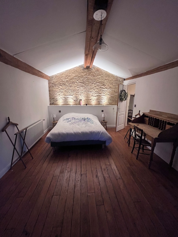 A bedroom with a sloped ceiling, wooden floor, a bed with a white and blue bedspread, a small table with chairs, and a wall-mounted light fixture.