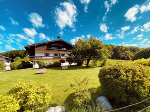 Un chalet à deux étages doté d’un balcon, entouré d’une végétation luxuriante et surplombant un ciel d’un bleu limpide.