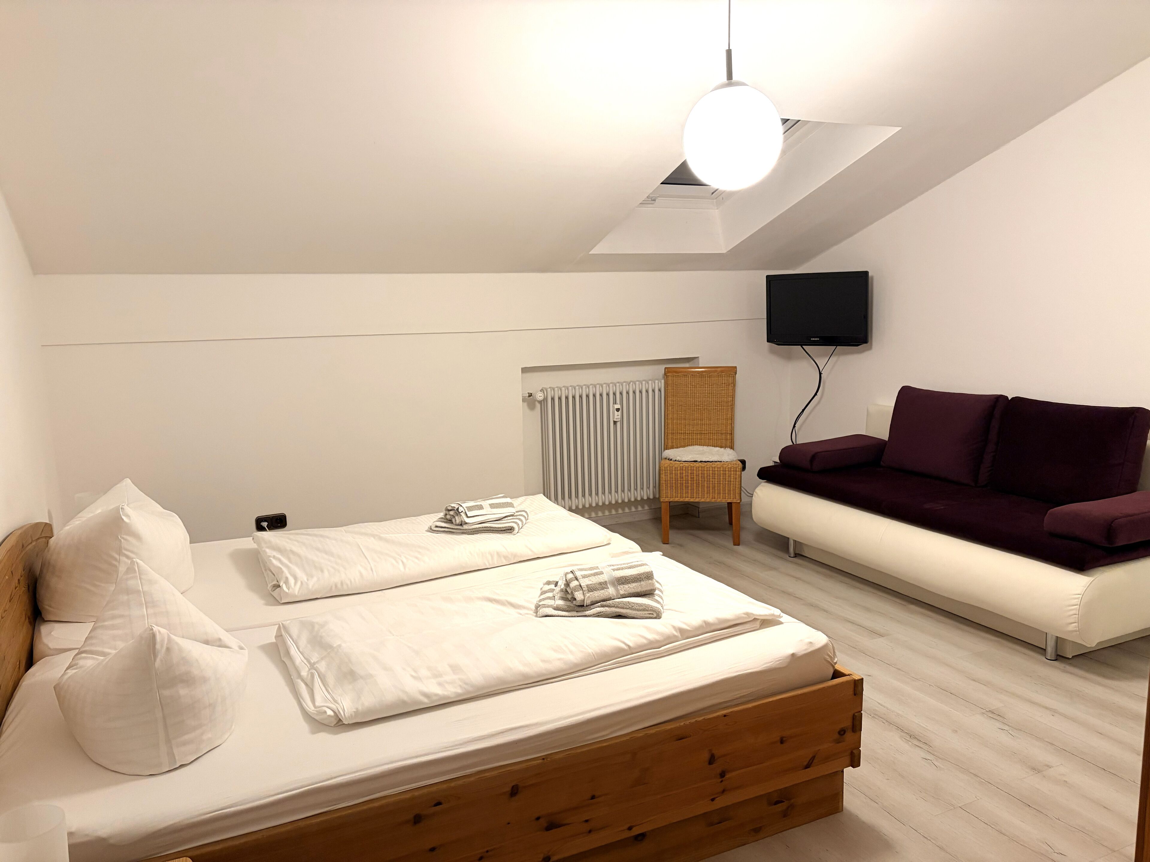 Ein modernes Schlafzimmer mit einem hölzernen Bett, einem weißen Sofa, einem Korbstuhl und einem flachbildfernseher an der Wand.