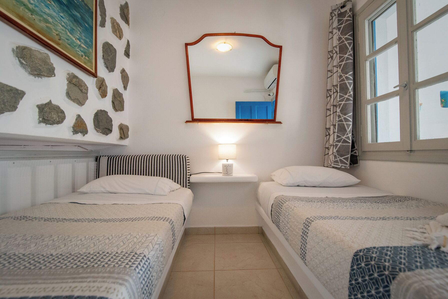 A room with two beds, a mirror, a small table with a lamp, and a window with patterned curtains.