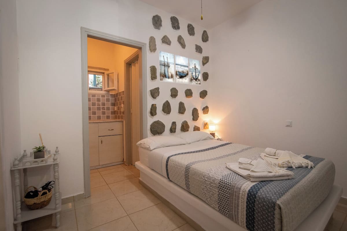 Une chambre comprenant un lit, une table de chevet, une étagère fixée au mur, un petit coin cuisine et une porte donnant sur une autre pièce.