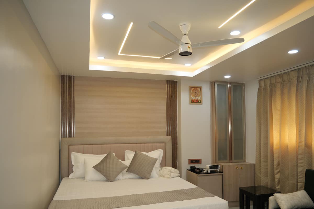 Una habitación de hotel moderna con una cama grande, un ventilador de techo, iluminación empotrada, una mesita de noche y un espejo, además de un ventanal con cortinas.
