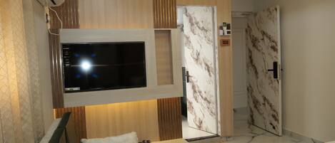 Una habitación de hotel moderna con televisor de pantalla plana montado en la pared, cama con cabecera, un pequeño rincón de descanso y baño con ducha de mármol.