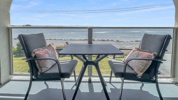 A balcony with two chairs and a table, overlooking a beach with a rocky shoreline. - Beachside Studio Steps from Casey’s Beach (Batehaven)