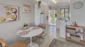 A modern kitchen with white appliances, a dining area with a round table and chairs, and a view of the outdoors through a glass door. - Beachside Studio Steps from Casey’s Beach (Batehaven)