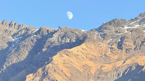 Paisaje montañoso con una luna en el cielo y un edificio de madera con techo de láminas de metal.
