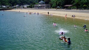 A beach with people swimming and playing in the water, a sandy shore, and a building in the background. - tabiyadoyobuko (Karatsu)