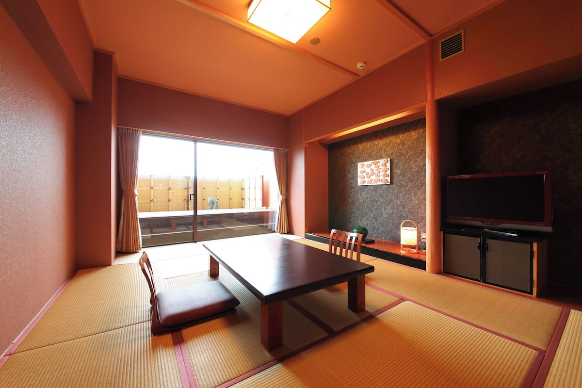 A traditional Japanese room with tatami flooring, a low table, chairs, a television, and a sliding door leading to a balcony.