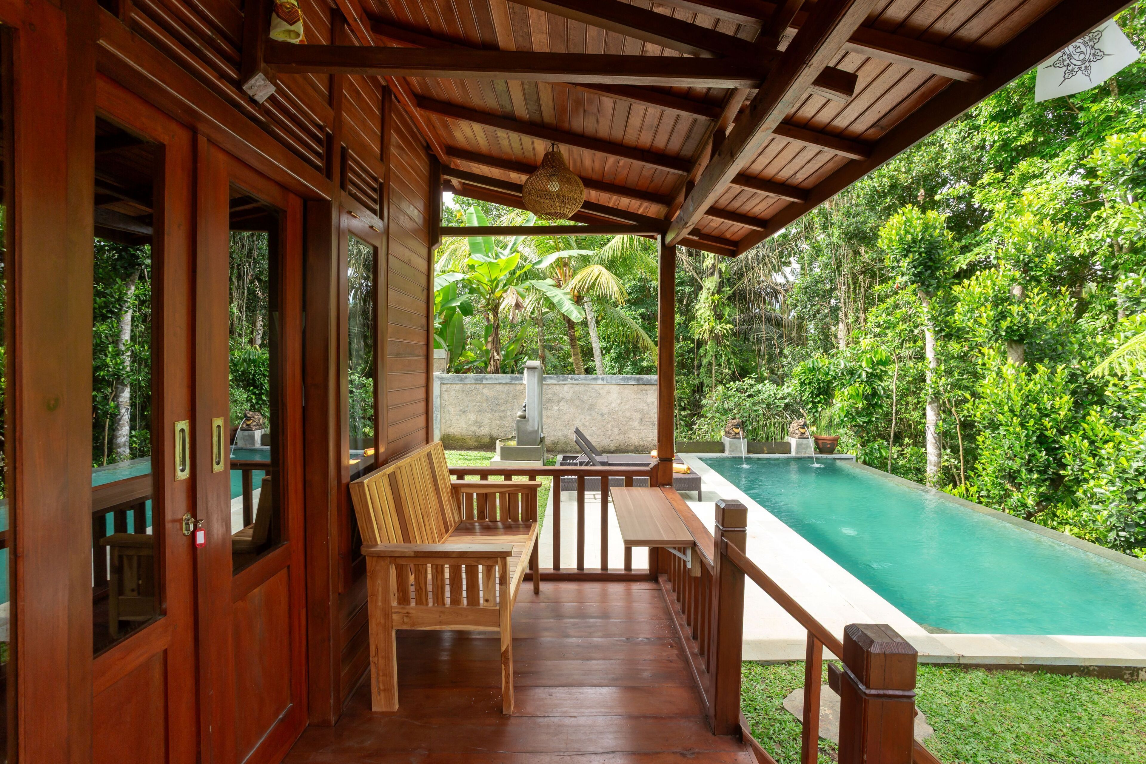 A wooden deck with a pool, wooden furniture, and a view of lush greenery.