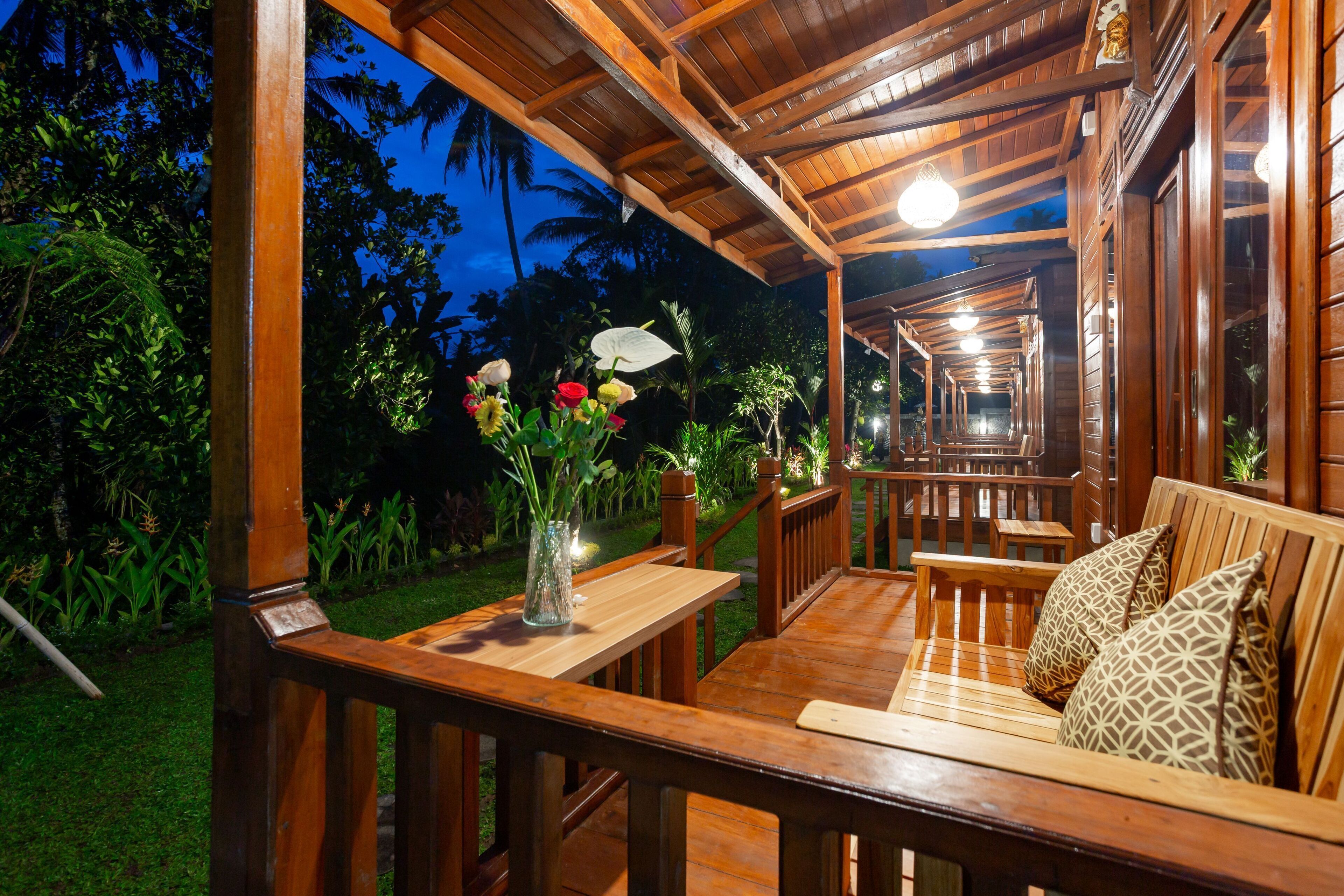 An outdoor wooden deck with a wooden bench and a table, surrounded by lush greenery and palm trees.