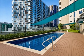 Ein Pool auf dem Dach mit einer Holzterrasse, umgeben von modernen Gebäuden und einem klaren Himmel.