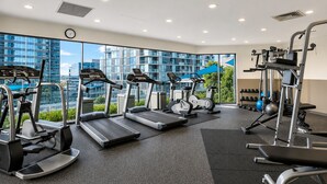 Une salle de sport équipée de tapis de course, de vélos elliptiques et d’appareils de musculation, de grandes fenêtres offrant une vue sur la ville et d’une horloge au mur.