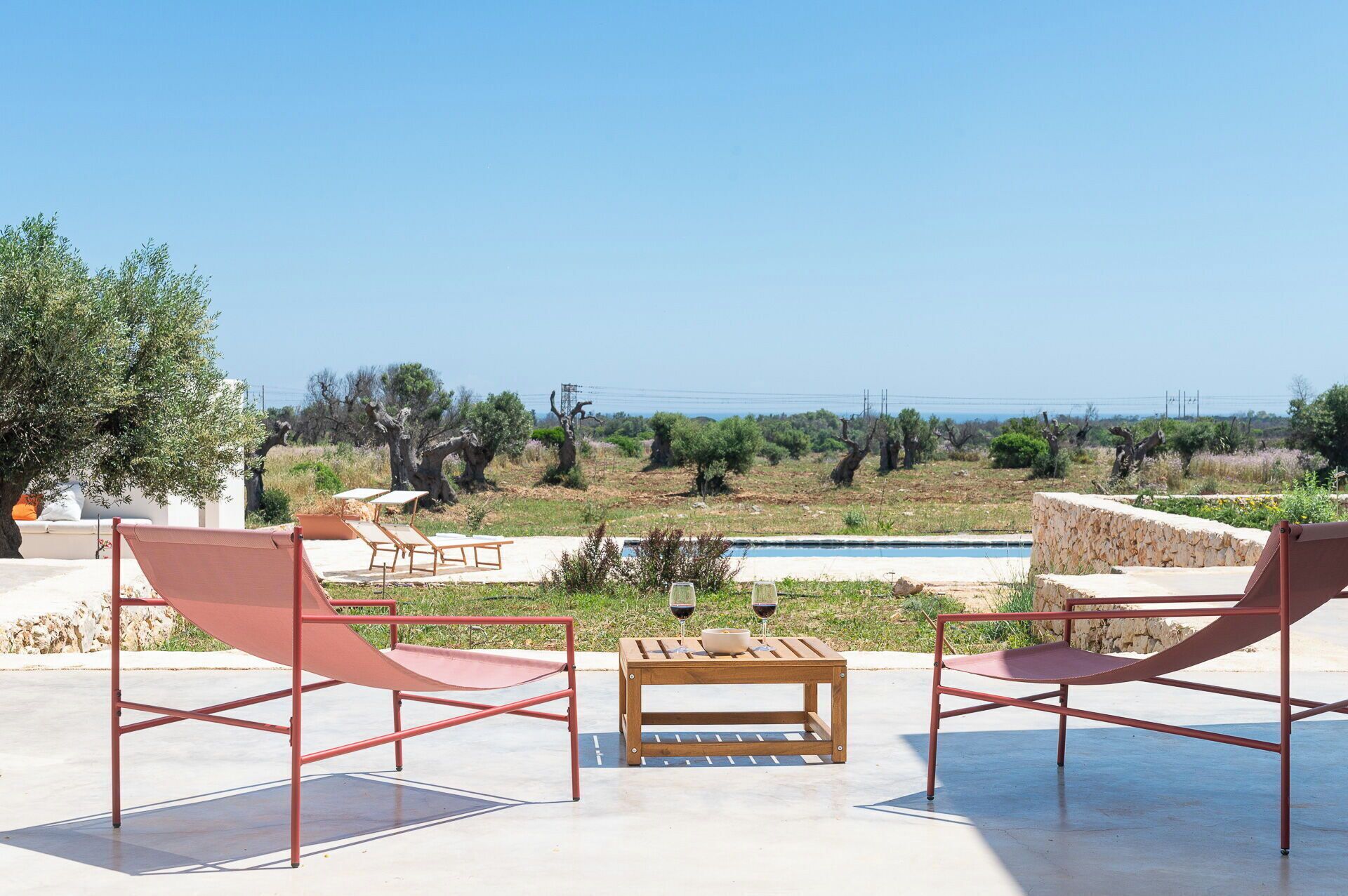 A patio with two red chairs and a wooden table, overlooking a swimming pool and a natural landscape.