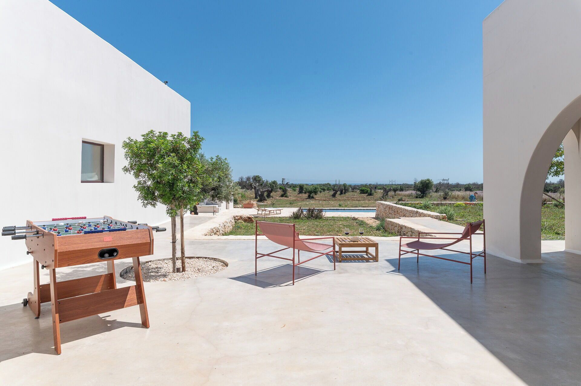 Eine Terrasse mit Tischfußball, zwei roten Stühlen, einem kleinen Tisch mit Weingläsern und Blick auf Pool und Gartenlandschaft.