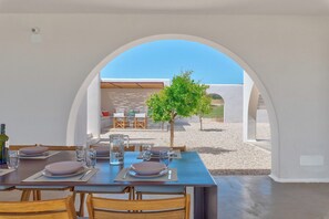Un coin repas avec une table dressée pour quatre personnes, un petit arbre et une vue dégagée sur l’extérieur, visible à travers une arche.