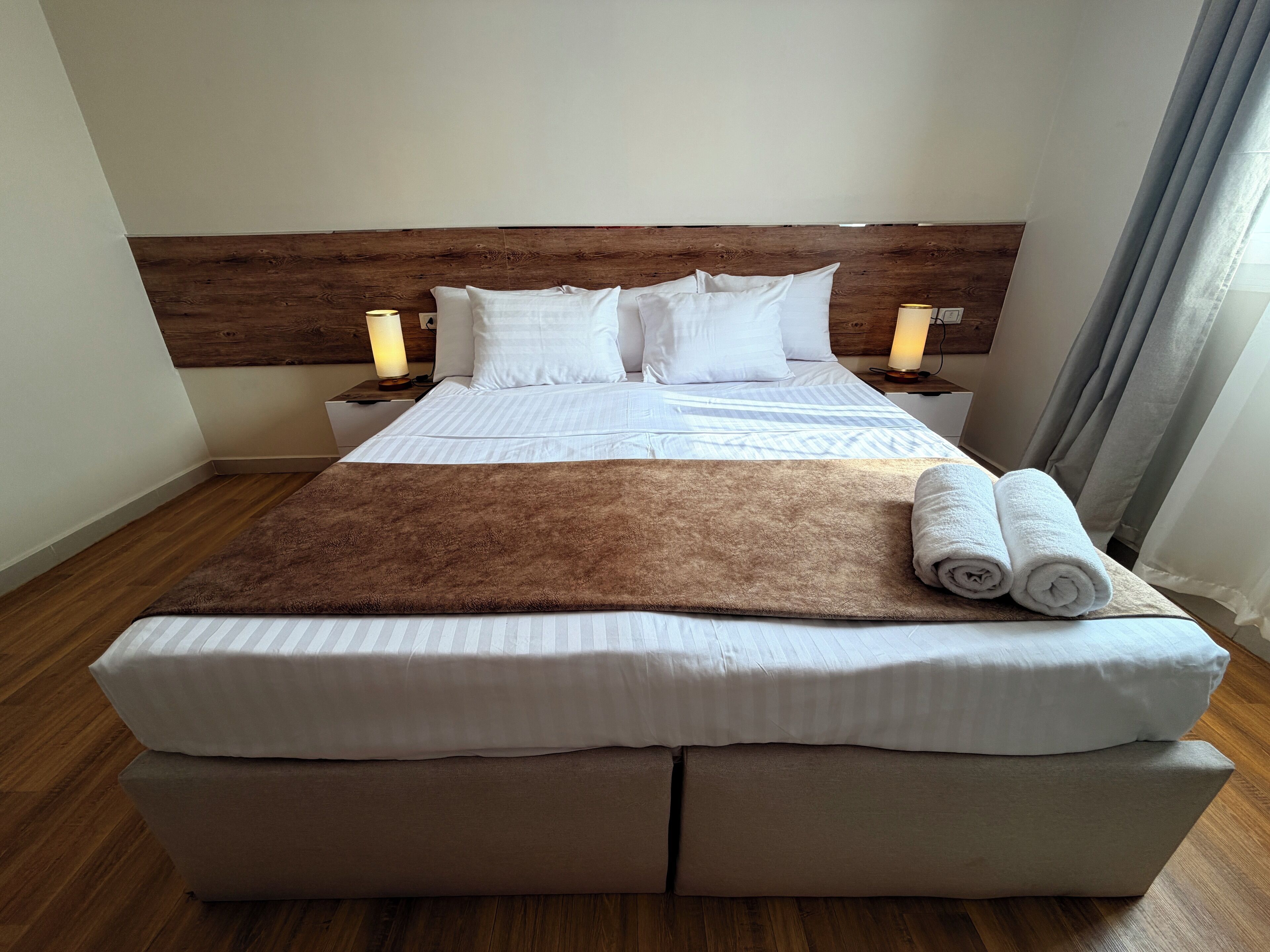 Een keurig opgemaakt bed met witte lakens, een bruine plaid en opgerolde handdoeken. Twee nachtkastjes met lampen, een houten hoofdbord en een houten vloer.