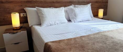 A neatly made bed with white linens and a brown throw blanket, flanked by two bedside tables with lamps, in a room with wooden headboard and a wooden floor.