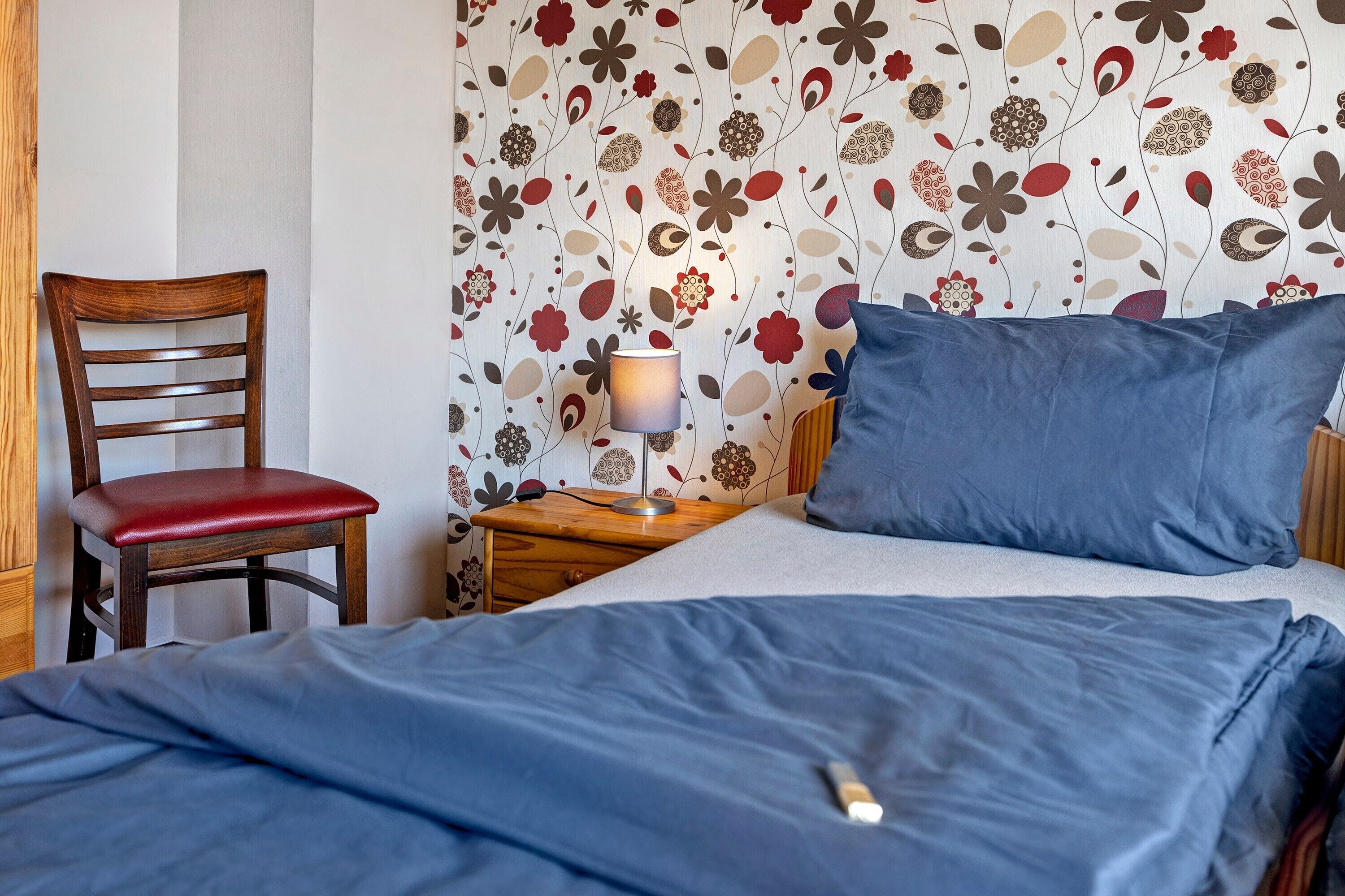Ein Schlafzimmer mit einem Bett, einem Sessel, einem Nachttisch mit Lampe und einer Wand mit Blumenmuster.