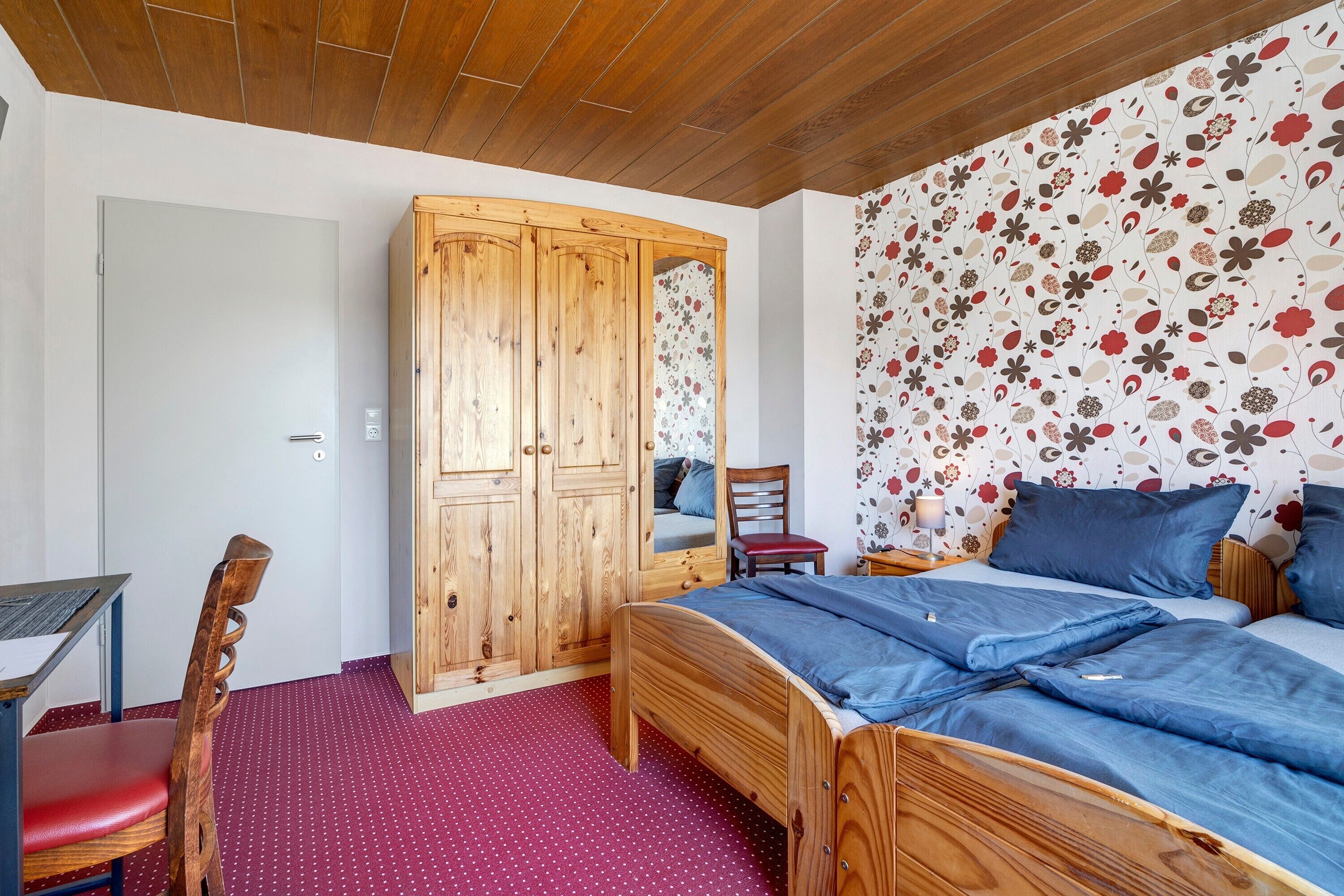 Une chambre avec une armoire en bois, un lit avec une literie bleue, une chaise rouge et un tapis à pois.