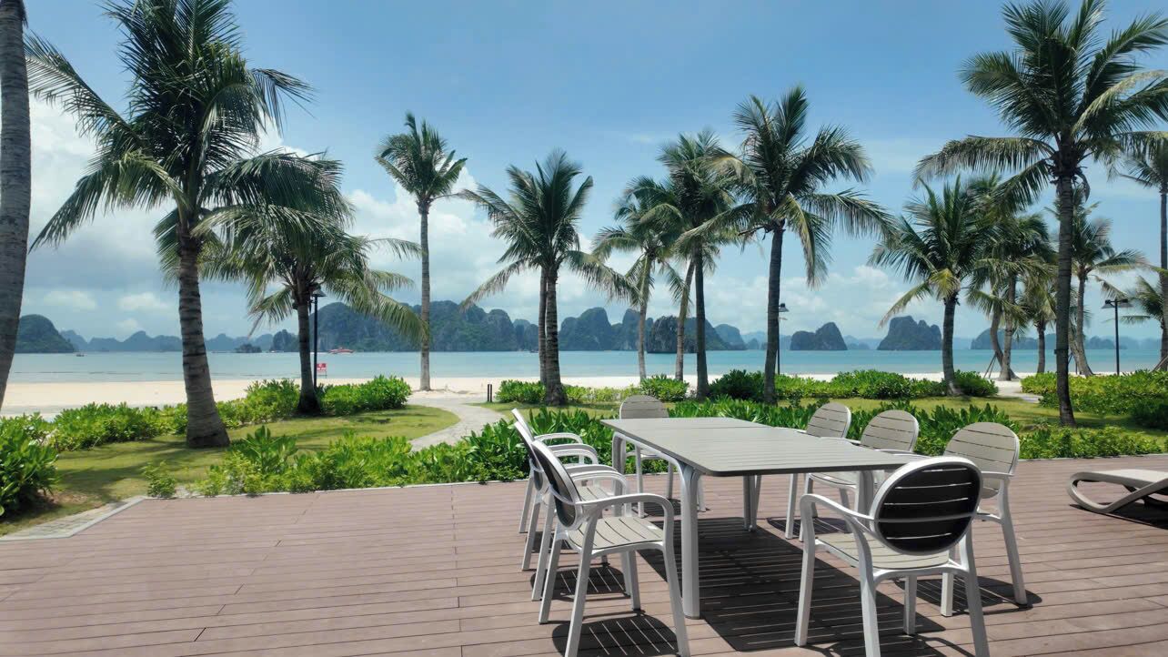 A deck with a table and chairs overlooking a beach with palm trees and distant mountains.