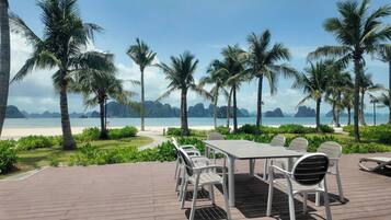 A deck with a table and chairs overlooking a beach with palm trees and distant mountains.