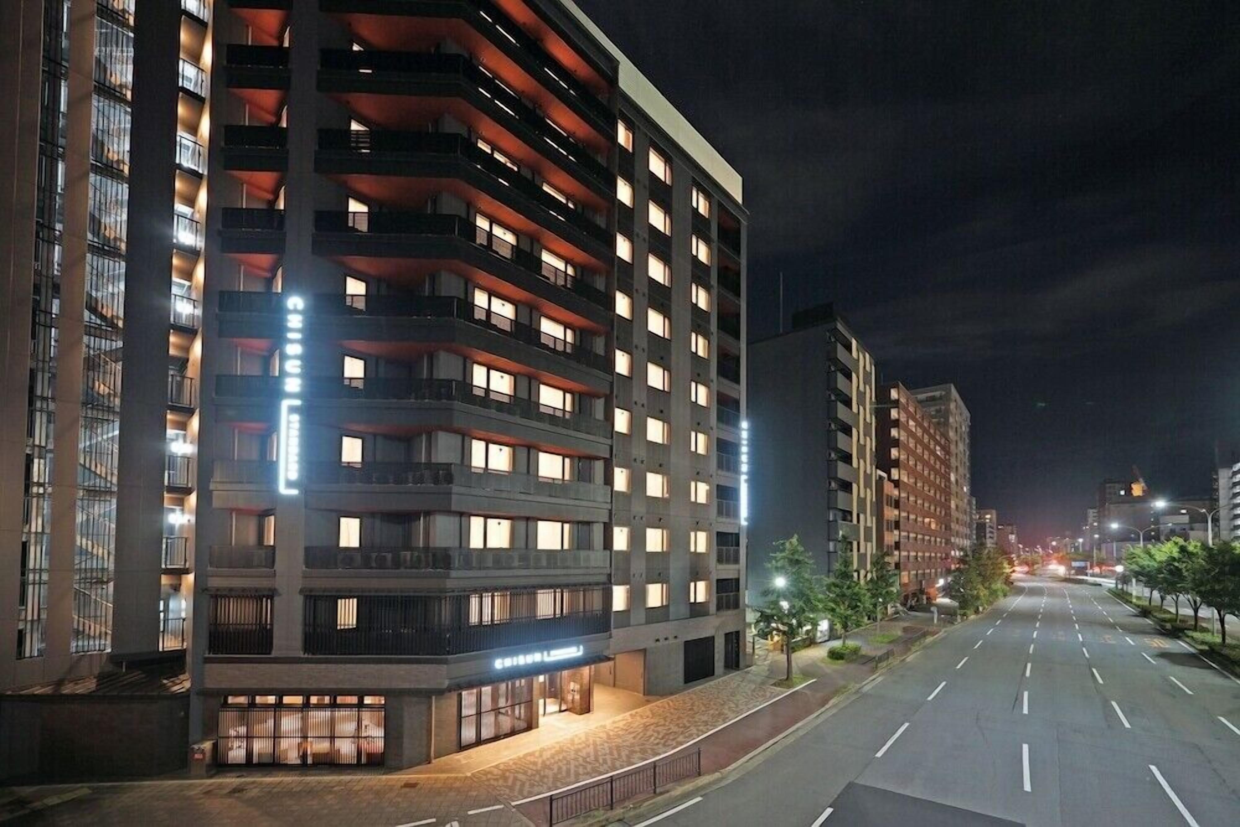 A multi-story hotel with illuminated windows and balconies, a clean street in front, and a clear night sky.