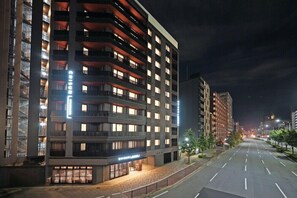Un hôtel à plusieurs étages, avec des fenêtres et des balcons illuminés, une rue propre devant et un ciel nocturne dégagé.