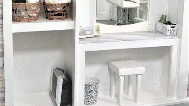 Een badkamer met een wit wastafelmeubel, een spiegel, twee rieten manden, een witte kruk en een kleine prullenbak.