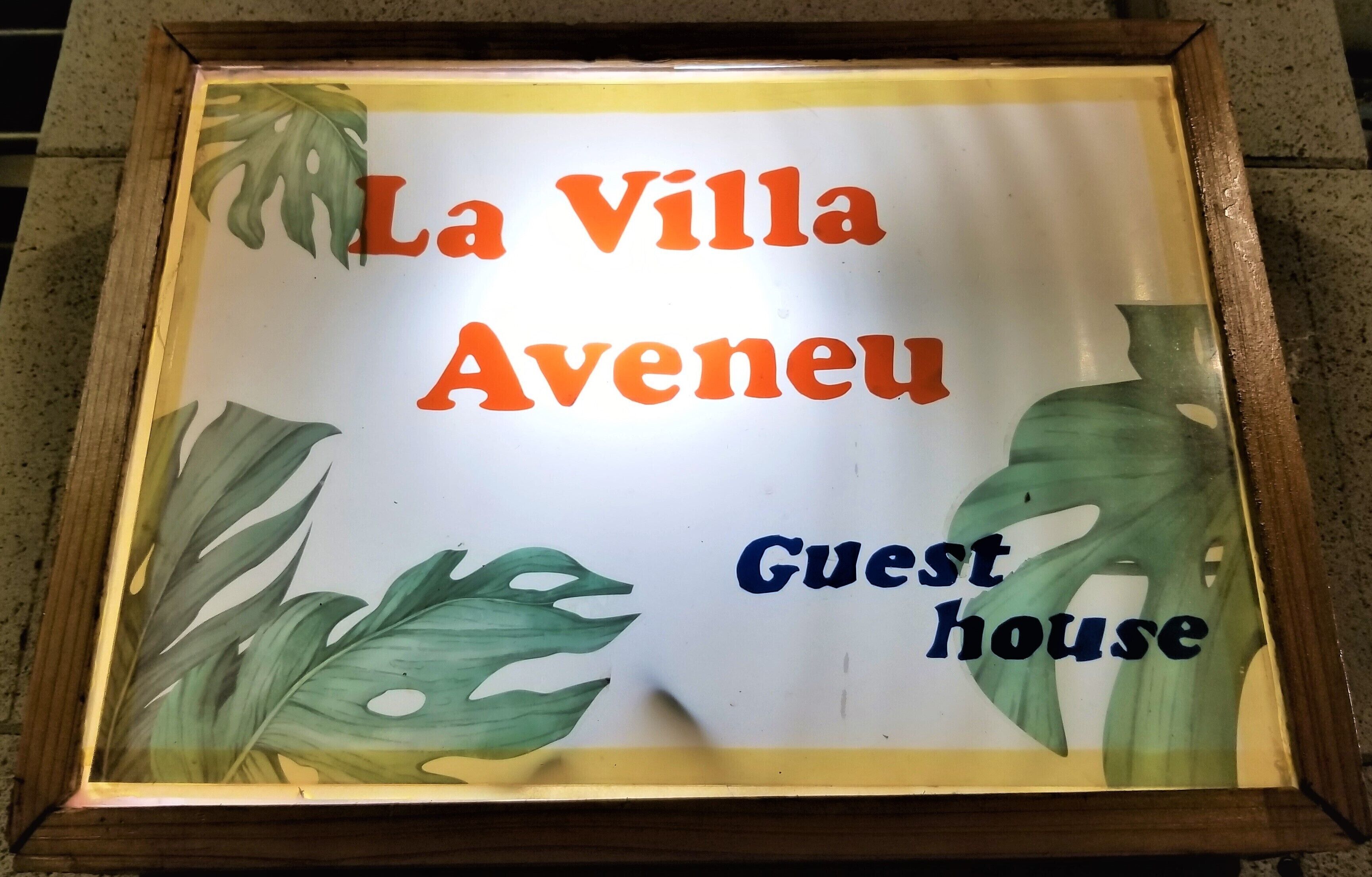 Un letrero en marco con la inscripción "La Villa Aveneu Guest House", rodeado de motivos con hojas tropicales.