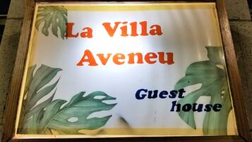 Un letrero en marco con la inscripción "La Villa Aveneu Guest House", rodeado de motivos con hojas tropicales.