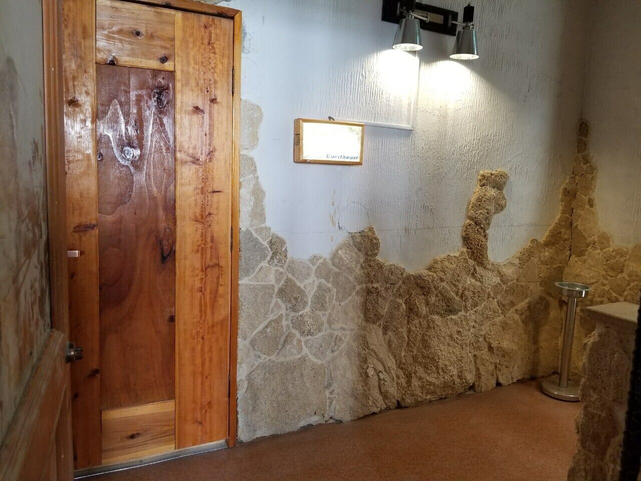 Una habitación con puerta de madera, pared de piedra, lámpara de metal y una pequeña mesa.