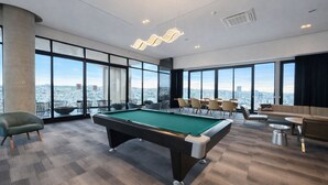 Un área de piscina interior moderna con mesa de billar, asientos y grandes ventanales que ofrecen vistas a la ciudad.