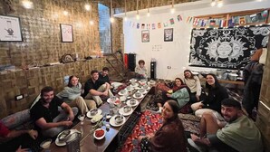 A group of people seated around a long table in a room with a wooden ceiling and walls, decorated with flags and artwork. - Yellowjackets Hotel Tosh (Tosh)