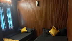 A room with two beds, each with a dark mattress and a yellow quilt. The walls have a vertical wooden panel design. - Olive Garden (Valparai)