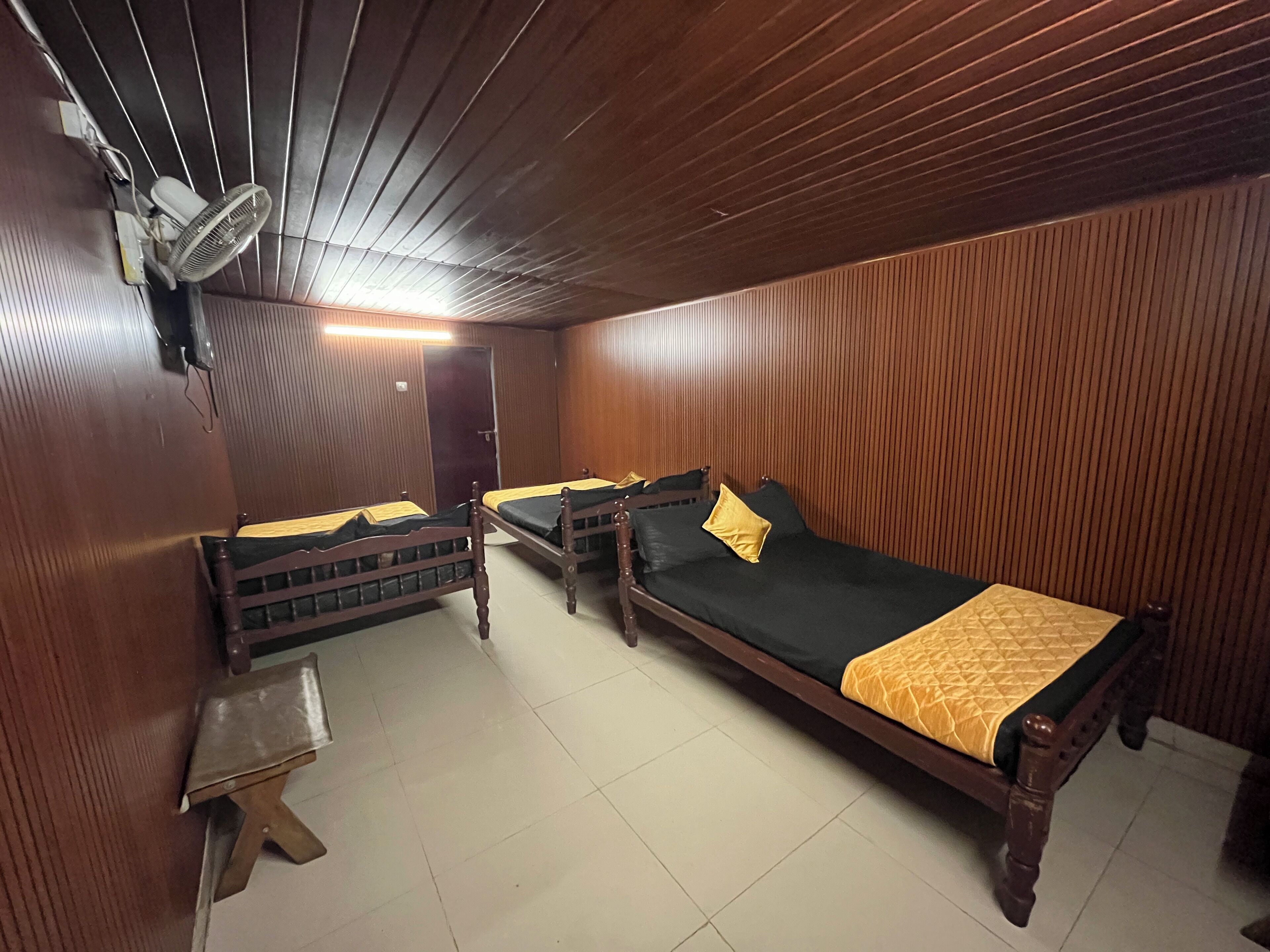 A room with wooden paneling, two single beds with yellow and black bedding, a small wooden table, and a wall-mounted fan.