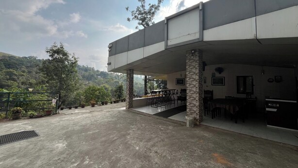 A covered outdoor seating area with tables and chairs, a staircase leading to an upper level, and a view of a hilly landscape. - Olive Garden (Valparai)