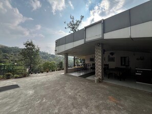 A covered outdoor seating area with tables and chairs, a staircase leading to an upper level, and a view of a hilly landscape. - Olive Garden (Valparai)