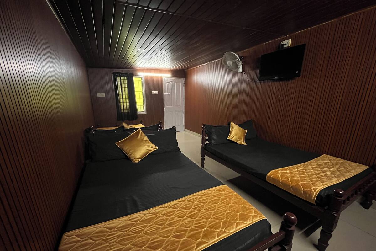 Une chambre avec deux lits, chacun doté d’un matelas noir et d’une couverture dorée. On y trouve également un ventilateur de plafond et un téléviseur fixé au mur.