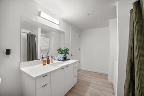 A modern bathroom with a large mirror, white cabinetry, a sink with toiletries, and a potted plant. - Walk to Downtown! Modern Riverfront Loft (West Sacramento)