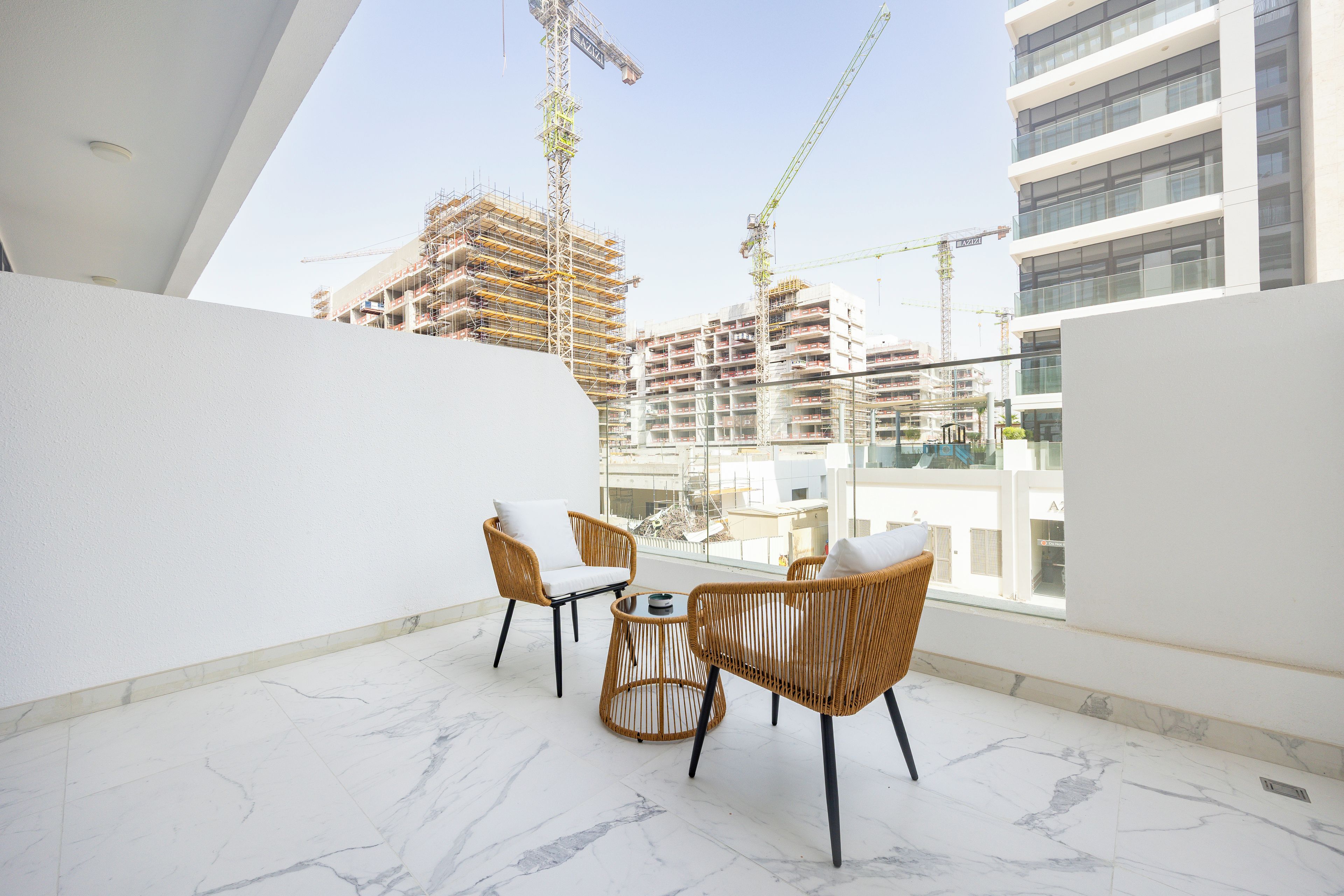 Ein Balkon mit Korbmöbeln und Marmorboden, mit Blick auf eine Baustelle mit Kränen.