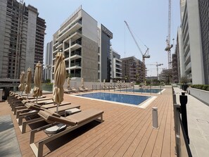 Uma área de piscina no terraço com espreguiçadeiras e guarda-sóis, rodeada por edifícios modernos e guindastes de construção.