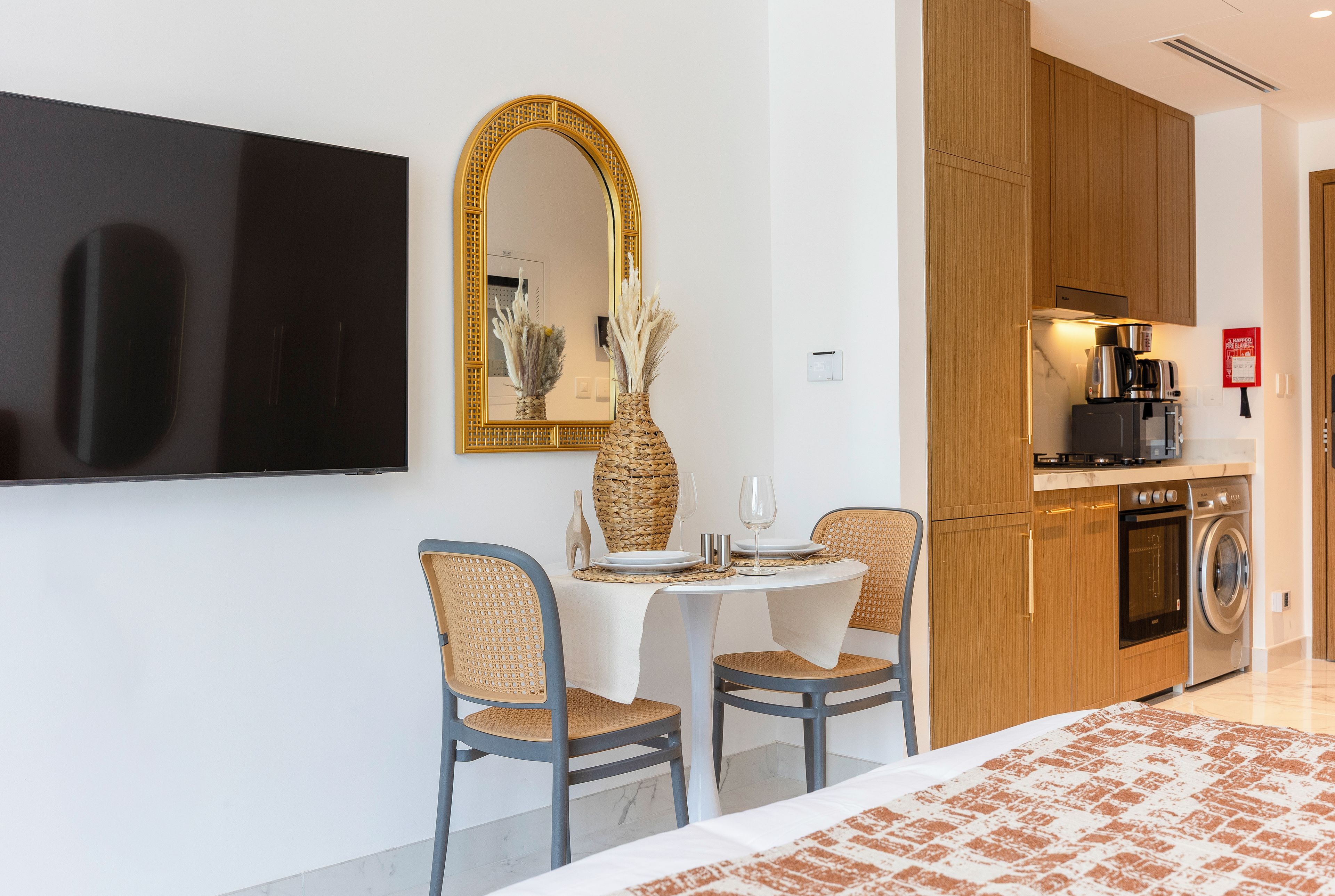 Una camera d'albergo moderna con TV a schermo piatto, un'area pranzo con due sedie e un tavolo, una cucina con macchina da caffè e lavatrice.